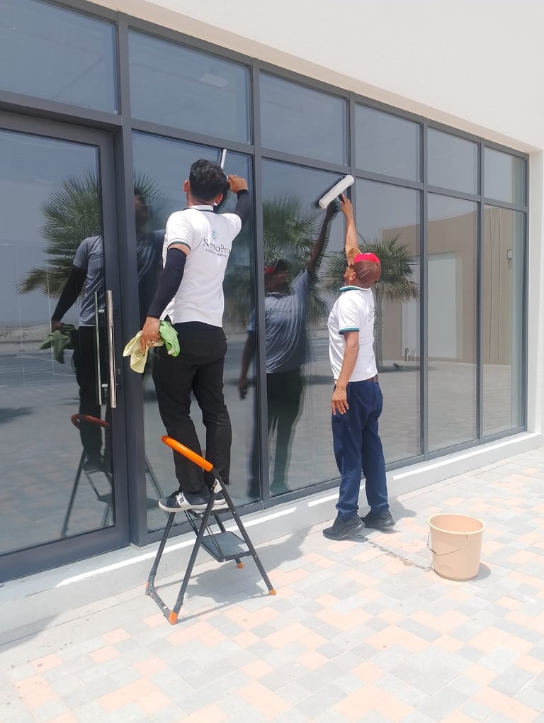 Two workers cleaning large glass windows on a modern building exterior with a ladder and cleaning tools