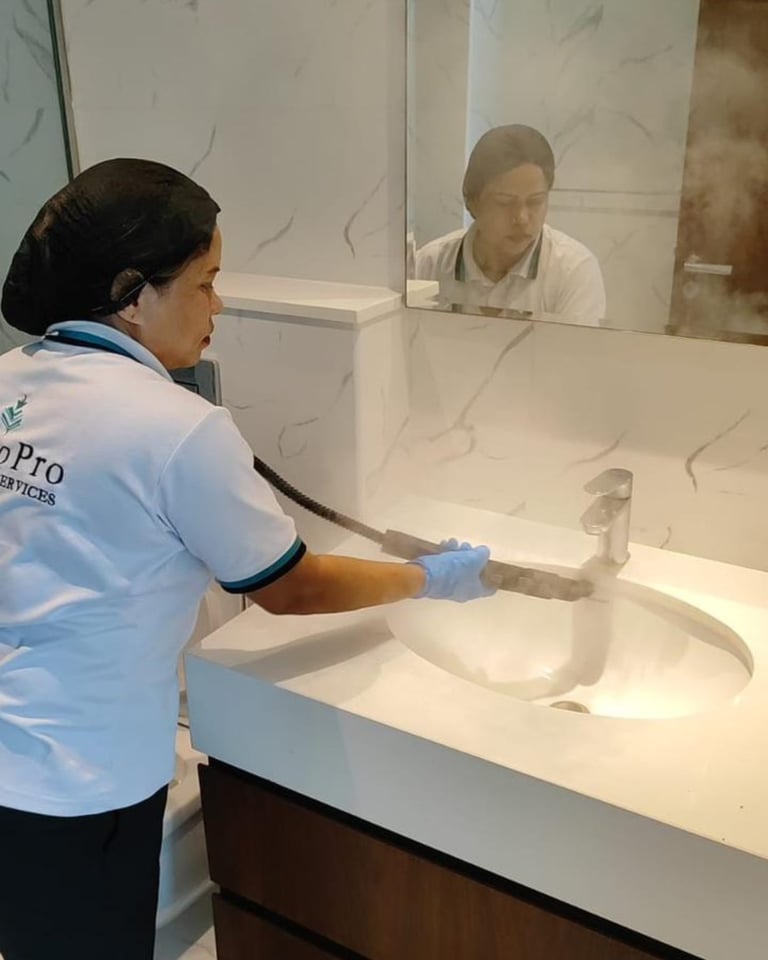 Cleaning professional in blue shirt cleaning bathroom sink counter with gloved hands while supervisor observes through glass partition